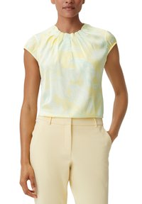 Femme portant un blouse sans manches à fleurs jaune clair et un pantalon taille haute jaune pâle avec poches, se tenant avec une main dans la poche.
