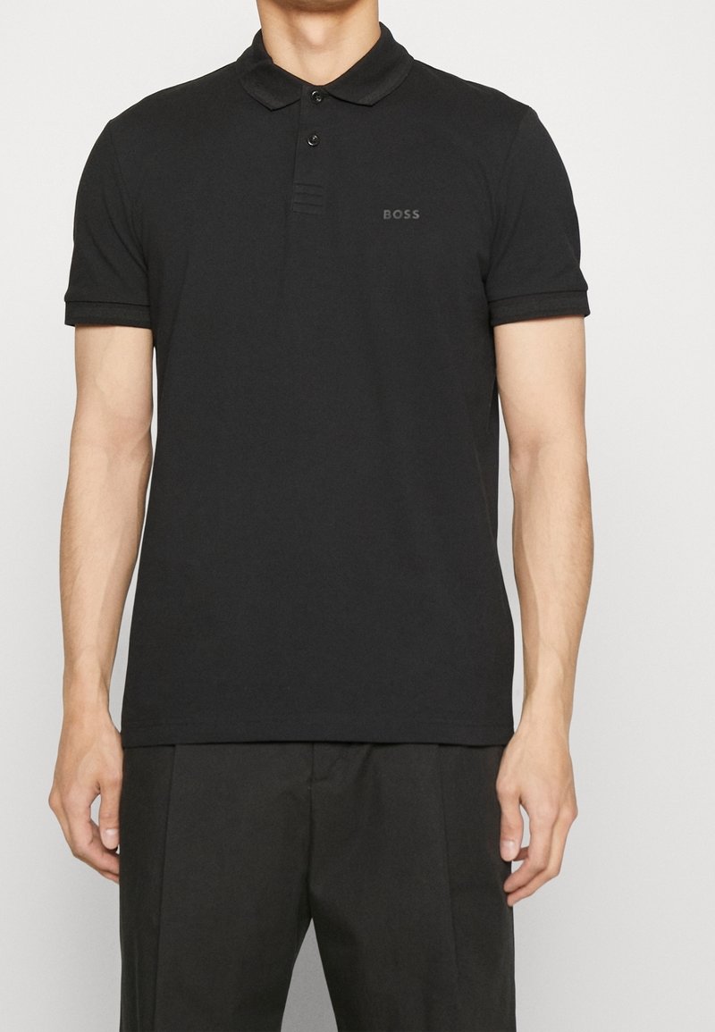 Homme portant un polo noir ajusté à manches courtes avec deux boutons et le logo "BOSS" sur la poitrine gauche, associé à un pantalon noir.