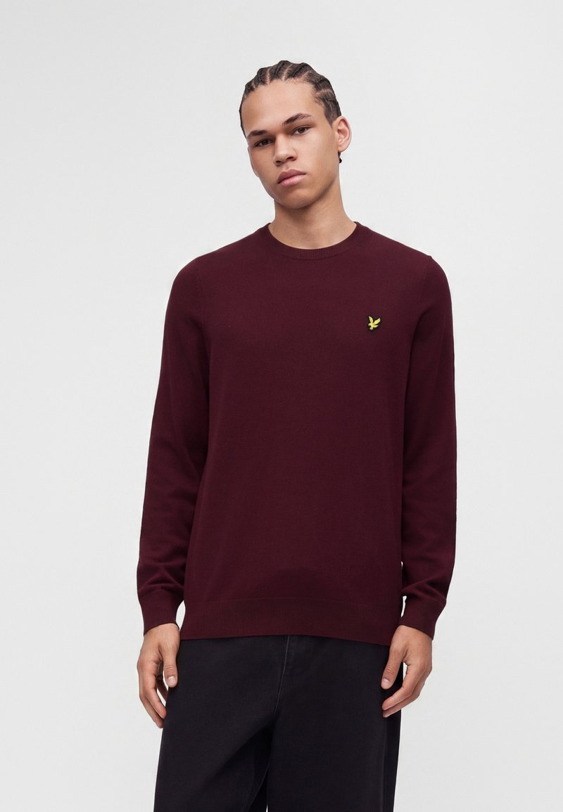Jeune homme aux cheveux tressés portant un pull uni bordeaux avec un logo d'oiseau jaune sur la poitrine, debout devant un fond blanc.
