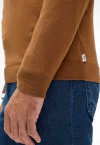 Bruine trui van gestructureerde stof, met ribgebreide boorden en zoom. Draagt over blauwe denim met een zichtbaar label op de zijnaad van de trui.