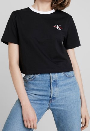 Kobieta ubrana w czarny krótki t-shirt z białym kołnierzykiem i logo Calvin Klein, w połączeniu z jasnoniebieskimi, wysokim stanem jeansami na szarym tle.