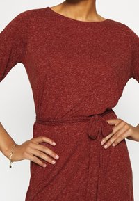 Frau in einem burgunderroten Kleid mit einem seitlichen Gürtel, die Hände in die Hüften gestützt, und einem goldenen Armband mit einem Vorhängeschloss-Anhänger am linken Handgelenk.