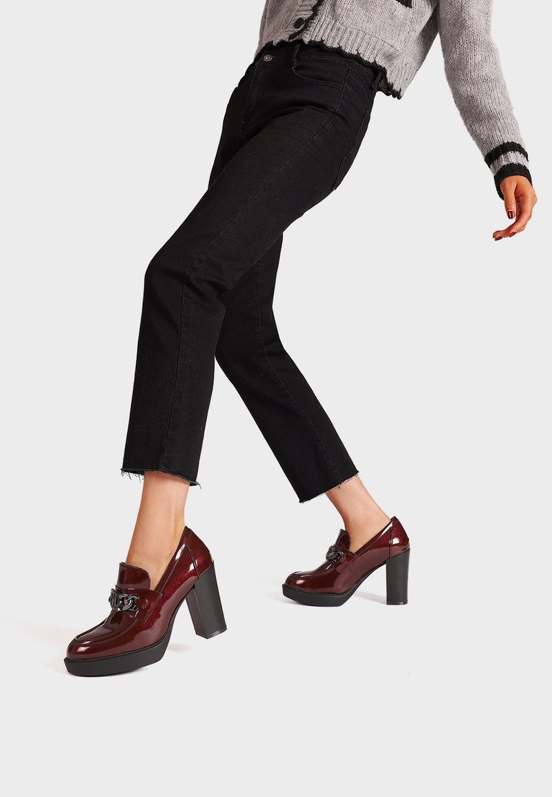 Scarpe platform bordeaux lucide con un tacco spesso e dettaglio decorativo a fibbia, indossate con jeans neri a vita alta e un maglione leggero.