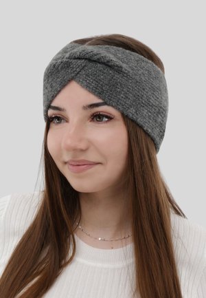 Giovane donna con lunghi capelli castani, che indossa un cerchietto grigio intrecciato in maglia e una maglietta bianca a coste, che sorride leggermente, sullo sfondo grigio chiaro.