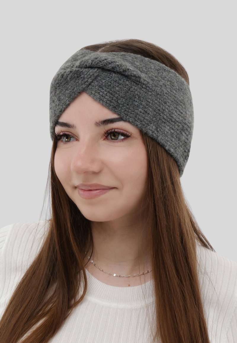 Young woman with long brown hair wearing a gray knitted twisted headband and a white ribbed top, smiling slightly, light gray background.