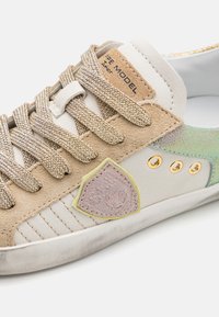 Beige suede and white leather sneaker with gold-accented eyelets, woven laces, and logo patch. Features textured iridescent side detail.