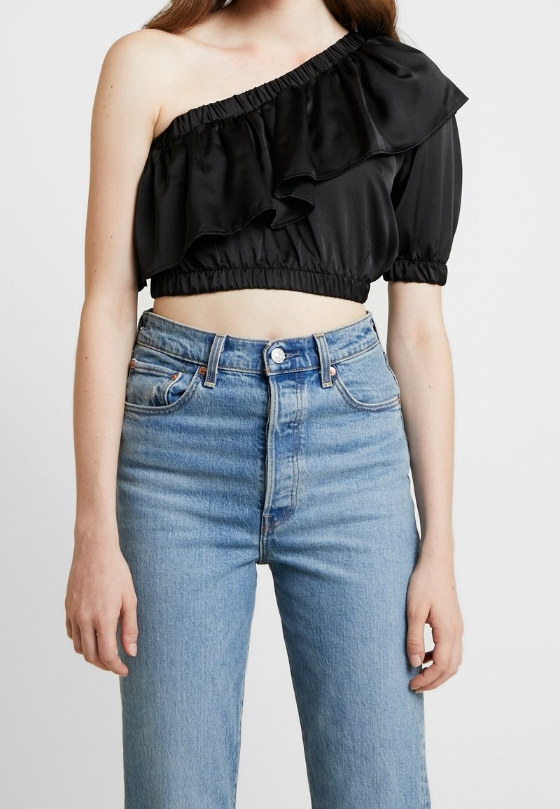 Svart satin cropped topp med en axel och volangdetaljer. Avslutad med ett elastiskt midjeband. Matchad med hög midja ljusblå jeans.