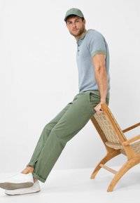 Polo di colore azzurro chiaro con motivo testurizzato e dettagli verdi, abbinato a pantaloni verde oliva e sneakers bianche, seduto su una sedia di legno.