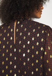 Blusa nera con un motivo di forme ovali rosse e dorate, con un'apertura a forma di chiave sul retro e maniche a palloncino.