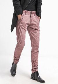 Rosa, figurnära byxor i ett texturerat tyg med rynkdetaljer vid anklarna och dekorativ sömnad på fickorna. Svarta ankelstövlar synliga.