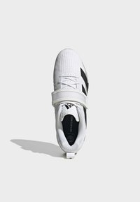 Bijela atletska cipela s teksturiranom tkaninom, crnim detaljima i širokim čičak trakama preko gornjeg dijela. Primijetite logotip na potplatu.