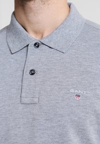 Szara koszulka polo wykonana z miękkiego materiału, z klasycznym kołnierzem, dwoma czarnymi guzikami oraz małym haftowanym logo na lewej piersi.