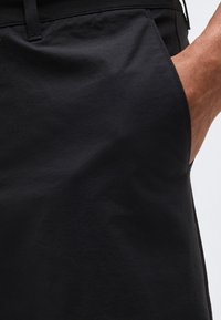 Κοντινή λήψη του χεριού ενός ατόμου που ακουμπάει μέσα στην εμπρόσθια τσέπη μαύρου παντελονιού με εμφανείς ραφές και θηλιές για ζώνη.