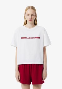 T-shirt bianca cropped con logo rosso "LACOSTE" e due strisce orizzontali rosse. Abbinata a pantaloni corti rossi, realizzati in morbido cotone.