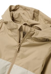 Beige Jacke mit Reißverschluss vorne und Kapuze, ausgestattet mit einem kontrastierenden cremefarbenen Streifen am unteren Rand. Glattes, leichtes, wasserabweisendes Material.