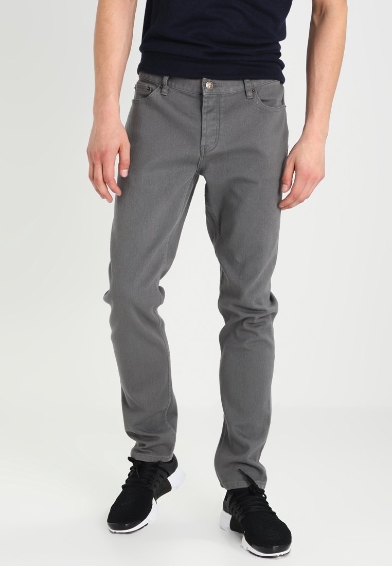 Homme portant un jean gris slim et des baskets noires, debout devant un fond uni, les mains détendues le long du corps.