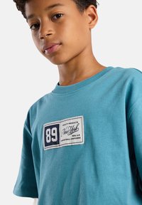 Turkoosi puuvillainen t-paita lyhyillä hihoilla, jossa on suorakulmainen logo-patch, jossa on tekstit "89" ja "New York" sinisellä ja valkoisella.