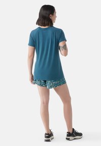 Türkisfarbenes Sport-T-Shirt mit kurzen Ärmeln, kombiniert mit gemusterten blauen Blumen-Shorts, von hinten betrachtet. Schwarze Sportschuhe sind getragen.
