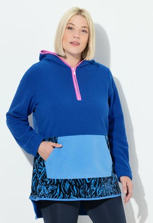 Kvinde med kort blondt hår bærer en blå hættetrøje med lyserød lynlås, lyseblå frontlomme og sort blomstermønster nær kanten.