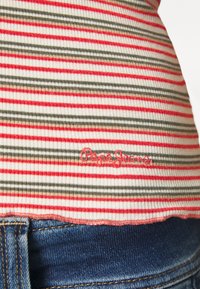Röd och grön randig ribbad tyg med vit bakgrund, med en broderad "Pepe Jeans"-logotyp nära fållen. Mörkblå denimmidja synlig.