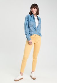 Chemise en denim à boutons de couleur bleu clair, associée à un pantalon jaune ajusté et des baskets blanches avec des semelles marron, se tenant devant un fond neutre.