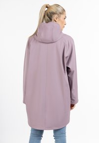 Hosszú, könnyű kabát puha levendula színben, kapucnival, sima textúrával és minimalista varrási részletekkel, farmernadrág felett viselve.