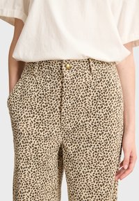 Pantaloni beige con stampa leopardata in tessuto strutturato, dotati di tasche laterali e chiusura con bottone frontale. Il design include piccole macchie scure sparse.