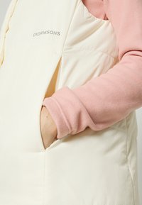 Colete acolchoado de cor clara feito de tecido suave, apresentando um bolso lateral e o nome da marca "Didriksons" impresso em cinza.