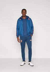 Μπλε φόρμα με ζακέτα με φερμουάρ και κάθετες ρίγες, συνοδευόμενη από αντίστοιχο παντελόνι. Διακοσμημένη με πορτοκαλί λογότυπα και λευκά αθλητικά παπούτσια.