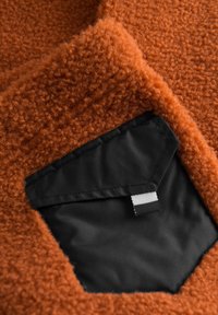 Rustfarvet, fuzzy stof med en blød tekstur, der har en kontrastfarvet sort, hexagonal lomme med en hvid stribet mærke detalje.