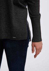 Grijze gebreide trui met geribbelde manchetten, voorzien van zijsplitten. Gecombineerd met donkere jeans, met een close-up van de arm en hand.