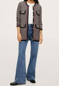 Gestrickte Strickjacke mit einem Marine- und Creme-Muster, ausgestattet mit goldenen Knöpfen und zwei Vordertaschen. Kombiniert mit ausgestellten blauen Jeans und schwarzen Schuhen.