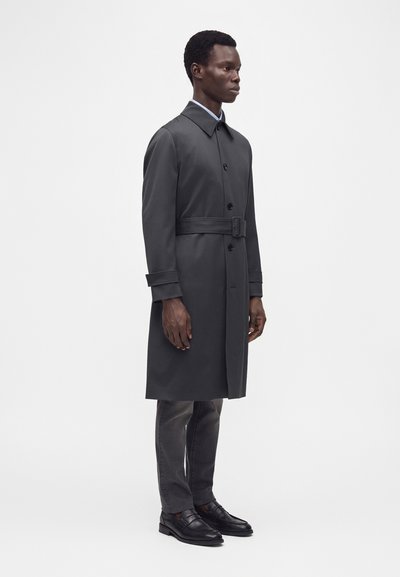 Homme debout portant un trench-coat gris foncé ceinturé, un pantalon foncé et des chaussures habillées noires sur un fond clair uni.