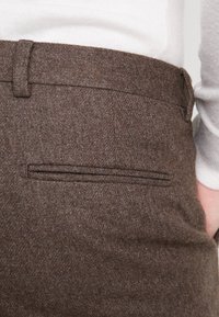 Close-up van bruine wollen broek met een paspel achterzak, gedragen met een witte gebreide trui.