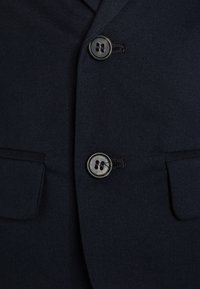 Mörkblå kostymjacka med detaljer som två svarta knappar, en slät tygtextur och två framfickor som är sydda med svarta trådkantningar.