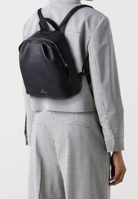 Zaino in pelle nera con forma arrotondata, chiusura con zip e spallacci regolabili. Dettagli in metallo dorato e superficie texturizzata.