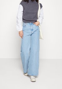 Helle blaue weit geschnittene Jeans mit hoher Taille, kombiniert mit einem dunkelgrauen, gerippten ärmellosen Oberteil und cremefarbenen Knöchelstiefeln.