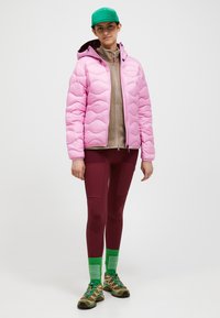 Giacca a vento imbottita rosa a trapunti, felpa leggera marrone chiaro con zip sotto, leggings bordeaux, berretto verde e calzini alla caviglia, scarpe sportive marroni e verdi.