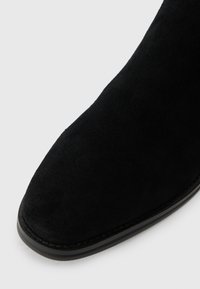 Czarne zamszowe botki za kostkę z ostro zakończonym noskiem, gładką fakturą oraz wąską czarną podeszwą z subtelnym detalem szwu.