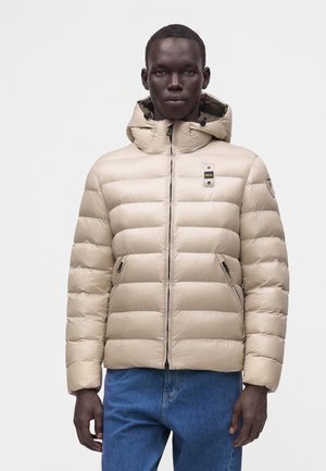 Mandlig model iført en lysebeige quiltet hættetrøjejakke med lynlåslommer og blå jeans, stående mod en ensfarvet hvid baggrund.