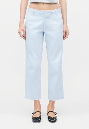 Ženska, oblečena v svetlo modre kratke hlače in črne balerinke s srebrnimi zakovicami, stoji ob belem ozadju.