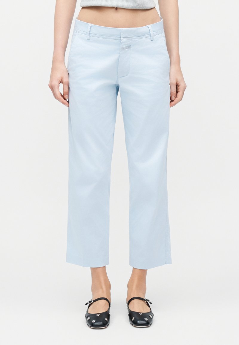 Γυναίκα που φοράει γαλάζια cropped παντελόνια και μαύρα φλατ παπούτσια με ασημένια διακοσμητικά, στέκεται μπροστά σε λευκό φόντο.