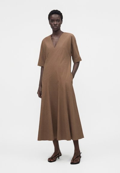 Γυναίκα που φοράει καφέ midi φόρεμα με μανίκια μέχρι τον αγκώνα και V-λαιμόκοψη, στέκεται με το ένα χέρι στην τσέπη, φοράει μαύρα σανδάλια με τακούνι.