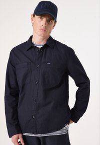 Navyblauwe overhemden met knopen en twee borstzakken, gemaakt van een gestructureerde stof. Draagt over een gestreepte grijze shirt, gecombineerd met een marineblauw pet.