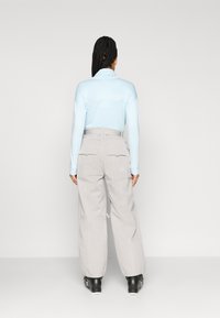 Camisa de manga longa azul clara com gola de fecho, combinada com umas calças largas cinza claro com bolsos laterais e punhos elásticos. Botas pretas.