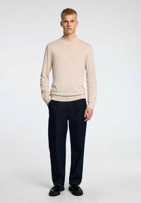Maglione lungo beige chiaro a maniche lunghe con collo a giro, abbinato a pantaloni plissettati blu scuro e scarpe di pelle nere. Tessuto semplice e strutturato.