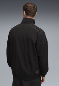 Sort zip-jakke med høj krave, der har røde og blå accenter, en glat tekstur og et broderet logo på den nederste ryg.