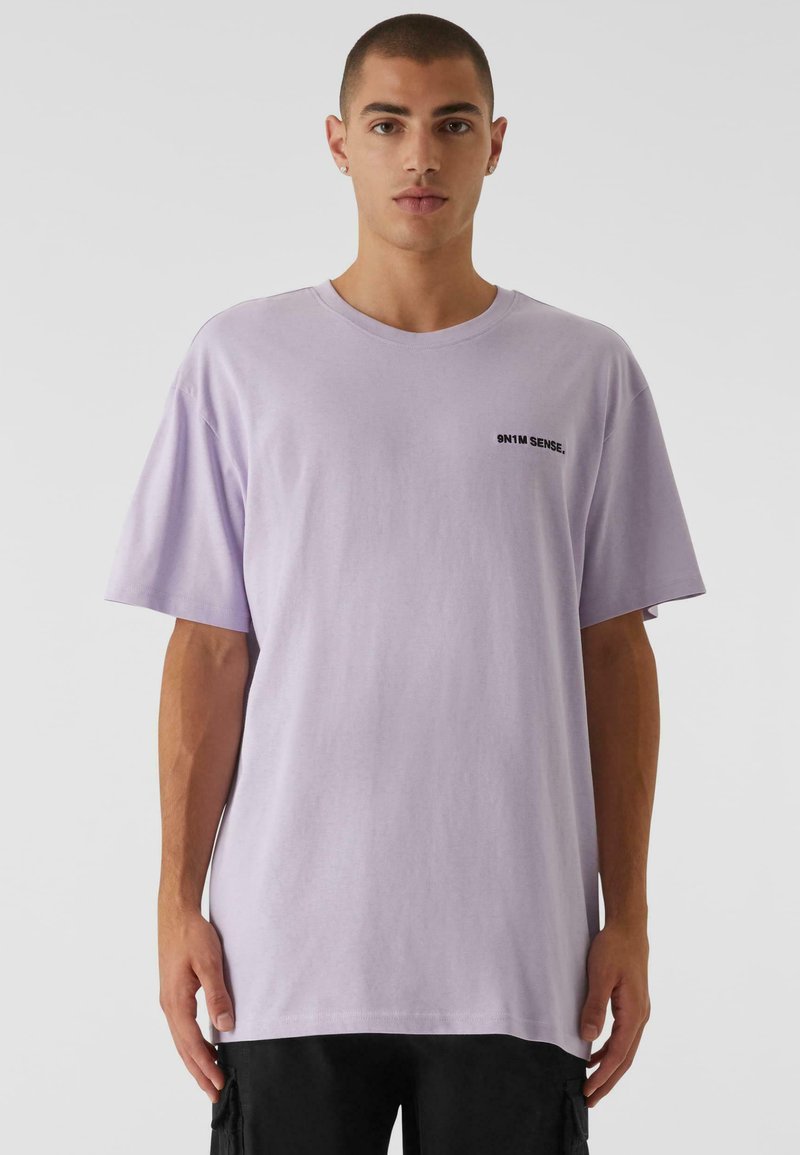 Camiseta de gran tamaño en color lavanda, hecha de algodón. Presenta un cuello redondo y mangas cortas, con el texto negro "9N1M SENSE." en el pecho.