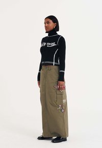 Schwarzer Strickrollkragenpullover mit weißen Details, kombiniert mit einem olivgrünen Cargo-Rock, der mit aufwändiger, mehrfarbiger Stickerei verziert ist. Schwarze Schuhe.