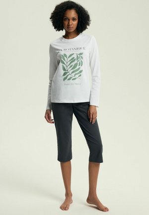 Woman standing barefoot wearing a white long-sleeve shirt with green leaf print and black cropped pants against a plain background.